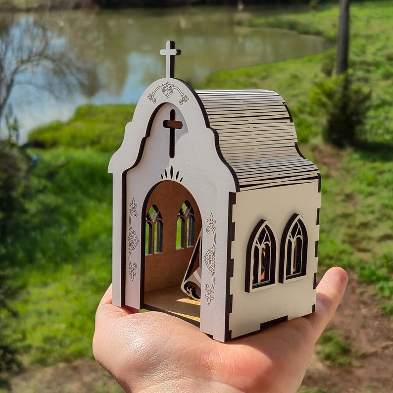 May include: A small, white, laser-cut wooden model of a church with dark brown edges. The structure features an arched entrance, a cross at the peak, and arched windows on the side. Intricate decorative patterns are etched onto the front facade.