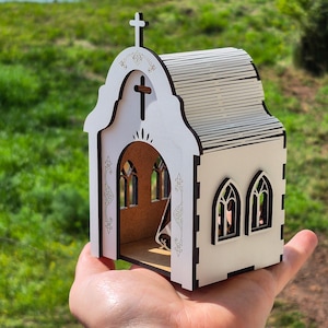 May include: A small, white, laser-cut wooden model of a church with a cross on the roof, arched windows, and intricate decorative engravings on the front facade. The structure features a brown interior base and dark edges from the cutting process.