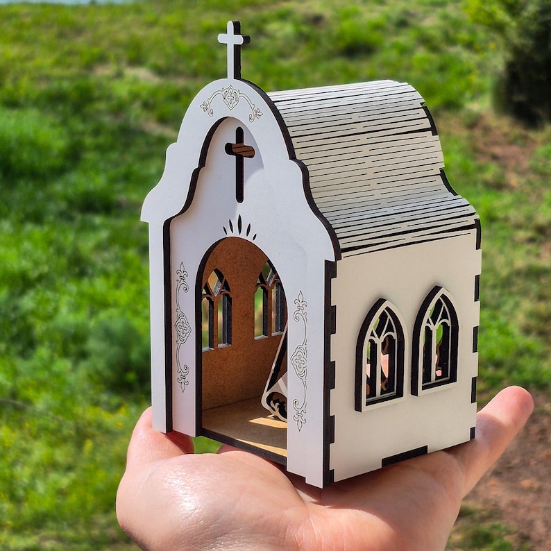 May include: A small, white, laser-cut wooden model of a church with a cross on the roof, arched windows, and intricate decorative engravings on the front facade. The structure features a brown interior base and dark edges from the cutting process.