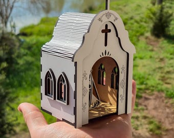 May include: A small, white, laser-cut wooden model of a church with dark edges. The structure features a peaked roof, a cross at the top, arched windows, and a large arched entrance. Intricate decorative scrollwork is etched along the front facade.