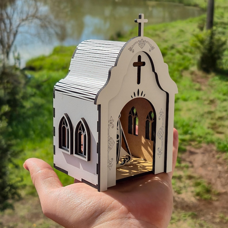 May include: A small, white, laser-cut wooden model of a church with dark edges. The structure features a peaked roof, a cross at the top, arched windows, and a large arched entrance. Intricate decorative scrollwork is etched along the front facade.