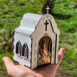 May include: A small, white, laser-cut wooden model of a church with dark brown edges. The structure features a curved front facade with a cross cutout, arched windows on the sides, a roof with horizontal lines, and a cross mounted on the peak.