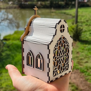 May include: A small, white, laser-cut wooden church model featuring a detailed arched stained-glass style window, two smaller arched side windows, a roof with horizontal slats, and a cross mounted on the peak of the front gable.