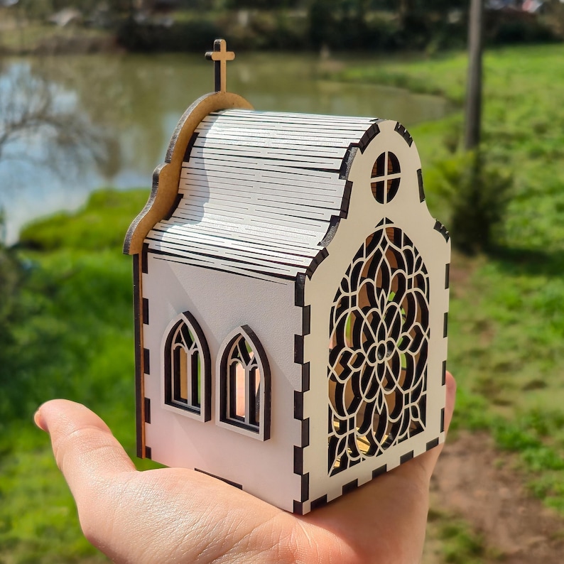 May include: A small, white, laser-cut wooden church model featuring a detailed arched stained-glass style window, two smaller arched side windows, a roof with horizontal slats, and a cross mounted on the peak of the front gable.