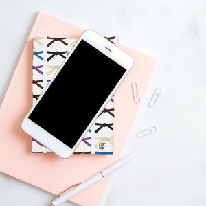 May include: A white smartphone with a black screen is lying on top of a pink notebook with a colorful pattern of martial arts belts. A white pen and three silver paper clips are also on the notebook.