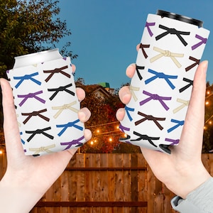 May include: Two white can coolers with a pattern of different colored martial arts belts. The belts are tied in a knot and are in the colors white, brown, black, purple, and blue.