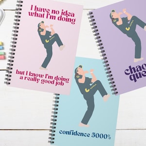 May include: Three spiral bound notebooks with different designs. The first notebook is pink with a cartoon of a person wearing a green and yellow uniform and the text "I have no idea what I'm doing but I know I'm doing a really good job". The second notebook is purple with a cartoon of a person wearing a green and yellow uniform and the text "chaotic queen". The third notebook is blue with a cartoon of a person wearing a green and yellow uniform and the text "confidence 5000%".