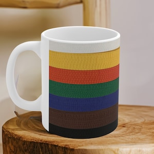Könnte beinhalten: Weiße Keramiktasse mit einem bunten Design aus übereinanderliegenden Kampfsportgürteln. Die Gürtel sind weiß, gelb, orange, grün, blau, braun und schwarz.