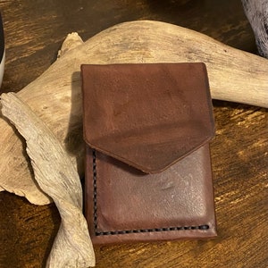 May include: A small, brown leather wallet with a flap closure. The wallet is hand-stitched with black thread along the edges. The leather has a slightly distressed appearance.