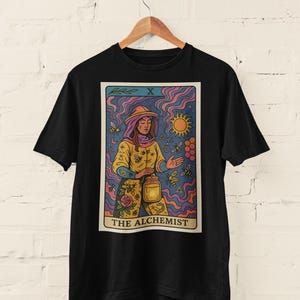 May include: Black t-shirt featuring a tarot card design with the text "THE ALCHEMIST." The card depicts a person in a yellow outfit with bees, a sun, and a honeycomb. The background has purple, blue, and pink wavy lines.