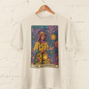 May include: Off-white t-shirt featuring a tarot card design. The card depicts a figure in a yellow outfit holding a lantern, surrounded by bees and a sun. The text "THE ALCHEMIST" is at the bottom of the card.