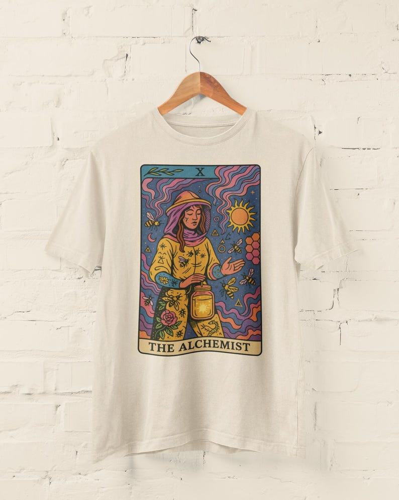 May include: Off-white t-shirt featuring a tarot card design. The card depicts a figure in a yellow outfit holding a lantern, surrounded by bees and a sun. The text "THE ALCHEMIST" is at the bottom of the card.
