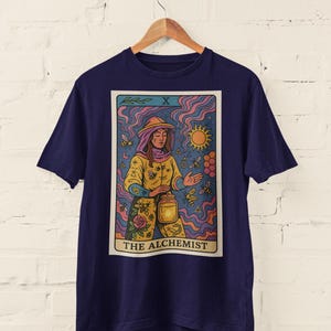 May include: Navy blue t-shirt featuring a tarot card design. The card depicts "The Alchemist" with a figure in yellow and orange, surrounded by bees and a sun. The design has a vintage, illustrated style.