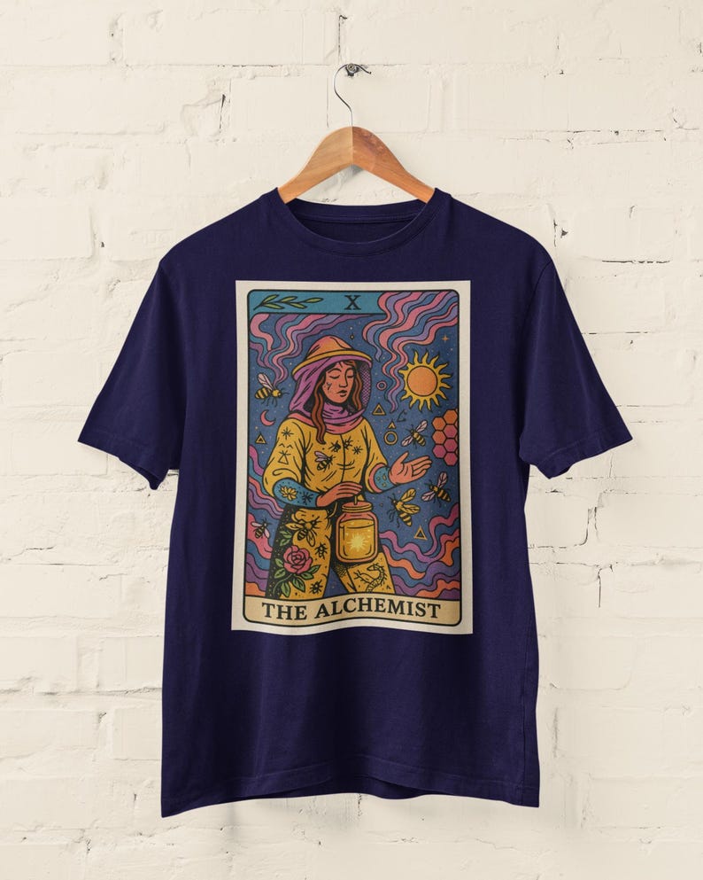 May include: Navy blue t-shirt featuring a tarot card design. The card depicts "The Alchemist" with a figure in yellow and orange, surrounded by bees and a sun. The design has a vintage, illustrated style.