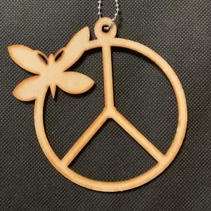 May include: A light-colored wooden ornament featuring a circular peace sign symbol with a dragonfly attached to the upper left side. The ornament is suspended by a silver-toned metal ball chain, showcasing a natural wood grain finish throughout.
