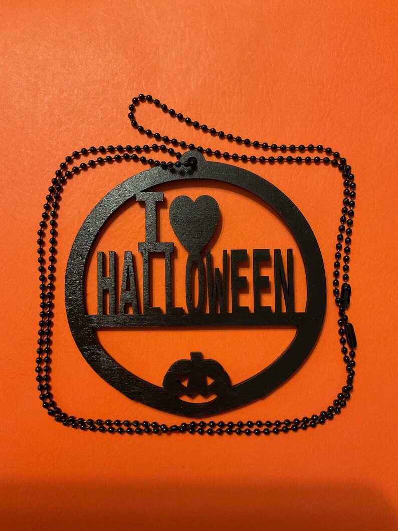 Op de afbeelding: Een zwart rond houten ornament met de tekst I HEART HALLOWEEN, waarbij een hartvorm het woord HEART vervangt en een uitgesneden pompoen met gezicht aan de onderkant. Het ornament is bevestigd aan een zwarte kralenketting.
