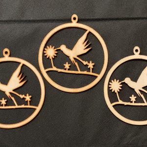 May include: Three circular light brown wooden ornaments, each featuring a laser-cut design of a hummingbird in flight approaching a flower. Each ornament has a small loop at the top for hanging and depicts the bird above a thin horizontal branch.