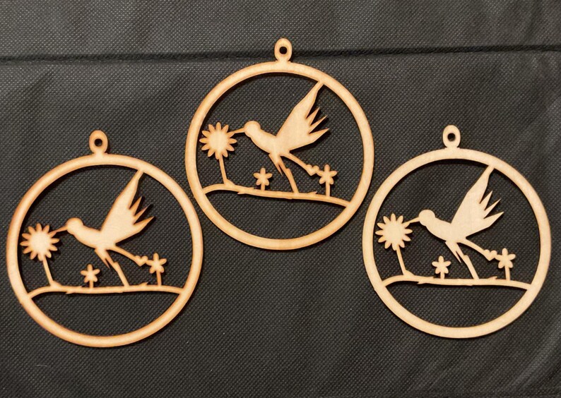 May include: Three circular light brown wooden ornaments, each featuring a laser-cut design of a hummingbird in flight approaching a flower. Each ornament has a small loop at the top for hanging and depicts the bird above a thin horizontal branch.