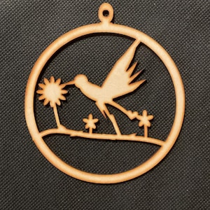 May include: A circular light brown wooden ornament featuring a laser-cut design of a bird in flight approaching a flower. The bird is positioned above a horizontal line with two smaller flowers below. A small loop at the top allows for hanging.
