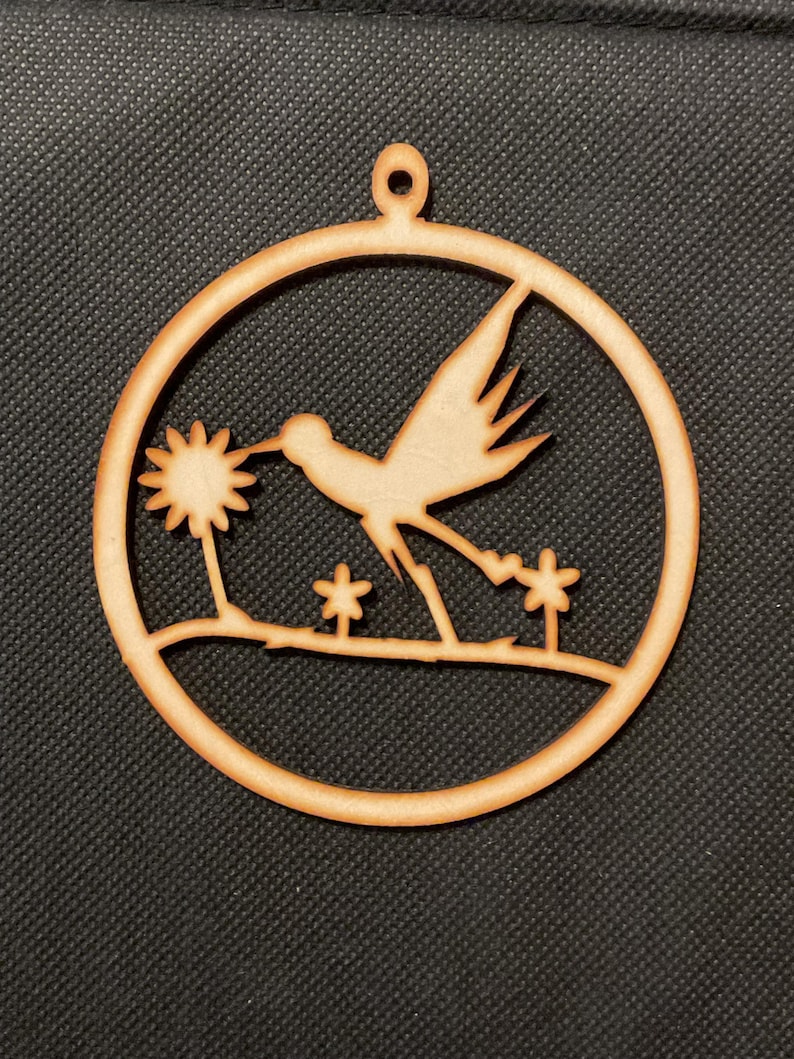 May include: A circular light brown wooden ornament featuring a laser-cut design of a bird in flight approaching a flower. The bird is positioned above a horizontal line with two smaller flowers below. A small loop at the top allows for hanging.
