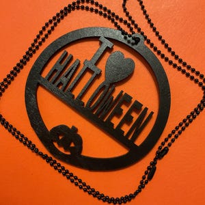 Op de afbeelding: Een zwarte ronde hanger aan een zwarte kralenketting. De hanger heeft een uitgesneden ontwerp met de tekst I HEART HALLOWEEN, waarbij een hartsymbool het woord HEART vervangt, en een klein pompoenicoontje aan de onderkant.