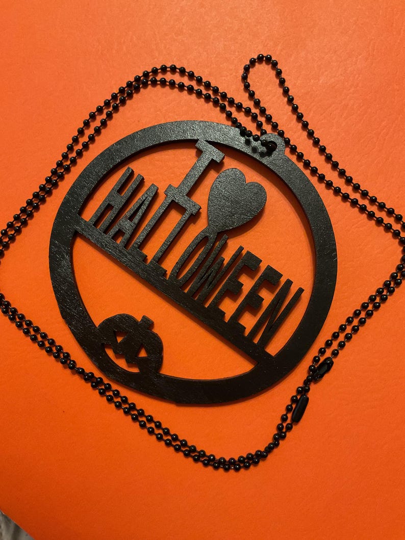 Op de afbeelding: Een zwarte ronde hanger aan een zwarte kralenketting. De hanger heeft een uitgesneden ontwerp met de tekst I HEART HALLOWEEN, waarbij een hartsymbool het woord HEART vervangt, en een klein pompoenicoontje aan de onderkant.