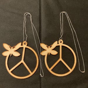 May include: Two light brown wooden peace sign ornaments, each featuring a small butterfly cutout attached to the upper left side of the circular frame. Each ornament includes a silver-toned metal ball chain for hanging purposes.