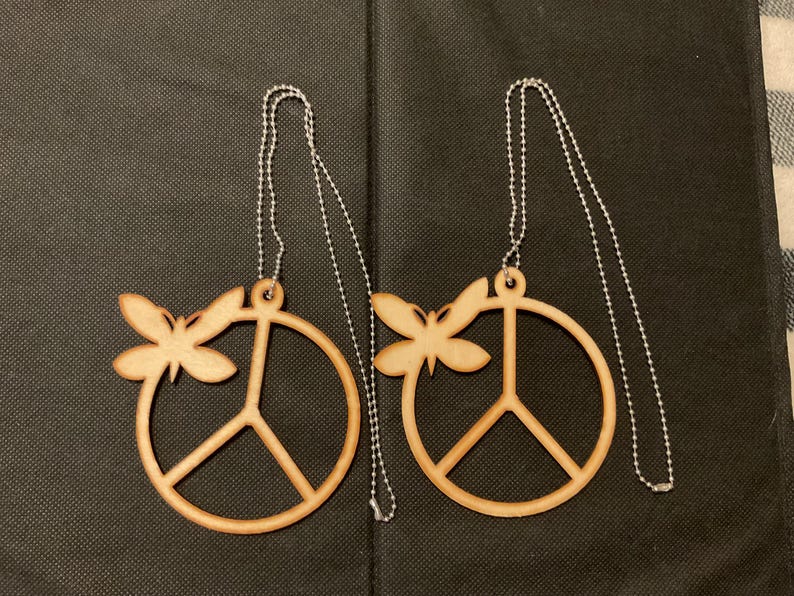 May include: Two light brown wooden peace sign ornaments, each featuring a small butterfly cutout attached to the upper left side of the circular frame. Each ornament includes a silver-toned metal ball chain for hanging purposes.