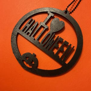 Op de afbeelding: Een cirkelvormig zwart ornament met de uitgesneden tekst I HEART HALLOWEEN, waarbij het woord HALLOWEEN onder een hartsymbool en de letter I is geplaatst, met een kleine pompoen-silhouet aan de onderkant, bevestigd aan een zwarte kogelketting.