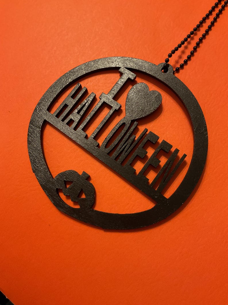 Op de afbeelding: Een cirkelvormig zwart ornament met de uitgesneden tekst I HEART HALLOWEEN, waarbij het woord HALLOWEEN onder een hartsymbool en de letter I is geplaatst, met een kleine pompoen-silhouet aan de onderkant, bevestigd aan een zwarte kogelketting.