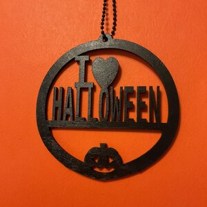 Op de afbeelding: Een zwart rond hangend ornament met de tekst I HEART HALLOWEEN, waarbij een hartsymbool de letter A vervangt en een uitgesneden pompoen aan de onderkant. Het ornament hangt aan een dunne zwarte kralenketting.