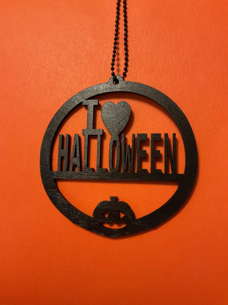 Op de afbeelding: Een zwart rond hangend ornament met de tekst I HEART HALLOWEEN, waarbij een hartsymbool de letter A vervangt en een uitgesneden pompoen aan de onderkant. Het ornament hangt aan een dunne zwarte kralenketting.