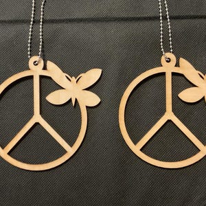 May include: Two light brown wooden ornaments, each featuring a circular peace sign symbol with a butterfly attached to the right side. Each ornament includes a small hole at the top with a silver-colored metal ball chain attached for hanging.
