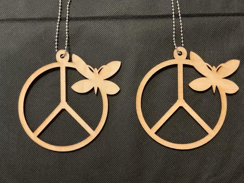 May include: Two light brown wooden ornaments, each featuring a circular peace sign symbol with a butterfly attached to the right side. Each ornament includes a small hole at the top with a silver-colored metal ball chain attached for hanging.