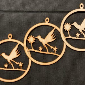 May include: Three circular light brown wooden ornaments, each featuring a laser-cut design of a hummingbird hovering near a flower. Each ornament has a small hole at the top for hanging and depicts the bird in flight above a thin branch.