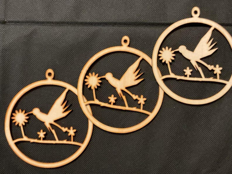 May include: Three circular light brown wooden ornaments, each featuring a laser-cut design of a hummingbird hovering near a flower. Each ornament has a small hole at the top for hanging and depicts the bird in flight above a thin branch.
