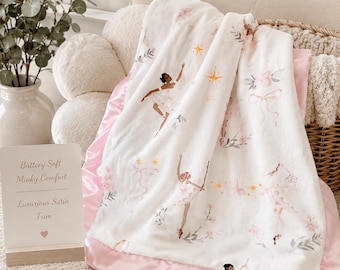 Könnte beinhalten: Eine weiße Minky-Decke mit einem Muster aus Ballerinas, rosa Blumen, Schleifen und goldenen Sternen, eingefasst mit einem rosa Satinband. Ein Schild daneben zeigt den Text: Buttery Soft Minky Comfort, Luxurious Satin Trim.