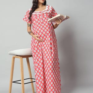 Puede incluir: Camisón de color coral con un estampado floral blanco y un ribete de encaje blanco alrededor del escote. El camisón de manga corta llega hasta el suelo. La persona sostiene un libro. Un taburete blanco y de madera está en el fondo.