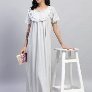 May include: A light gray nightgown with a ruffled neckline and short sleeves. The long, flowing gown has a buttoned bodice and a simple, elegant design. The woman is holding a book titled "It Ends With Us".