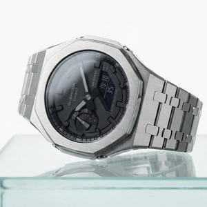 Può includere: Un orologio da polso argentato con quadrante grigio scuro e cinturino in metallo coordinato. L'orologio indica l'ora 10:10 e 35 secondi. L'orologio è appoggiato su una superficie di vetro trasparente.