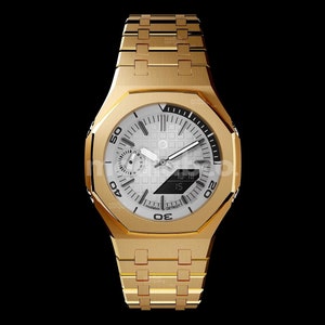 Op de afbeelding: Een gouden polshorloge met een witte wijzerplaat en zwarte accenten. Het horloge heeft een vierkante kast en een metalen band. De wijzerplaat van het horloge heeft een datumvenster en een chronograaf. Het horloge is gelabeld "MLC 346-C.CMYY.SY.DAW.MS.HS" en "BESPOKE STAINLESS STEEL 316L 48.5 x 45.4 x 11.9 mm HANDMADE IN DETROIT".