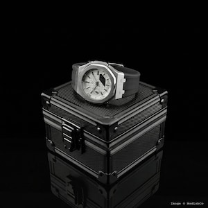 May include: A silver-toned wristwatch with a white face and black band rests on a black, textured box. The watch has a geometric design and visible time indicators. The box has a metallic appearance with silver trim and a clasp.