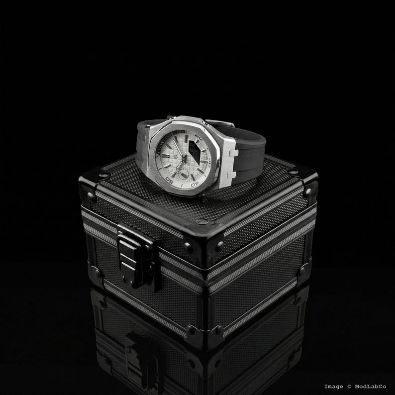 May include: A silver-toned wristwatch with a white face and black band rests on a black, textured box. The watch has a geometric design and visible time indicators. The box has a metallic appearance with silver trim and a clasp.