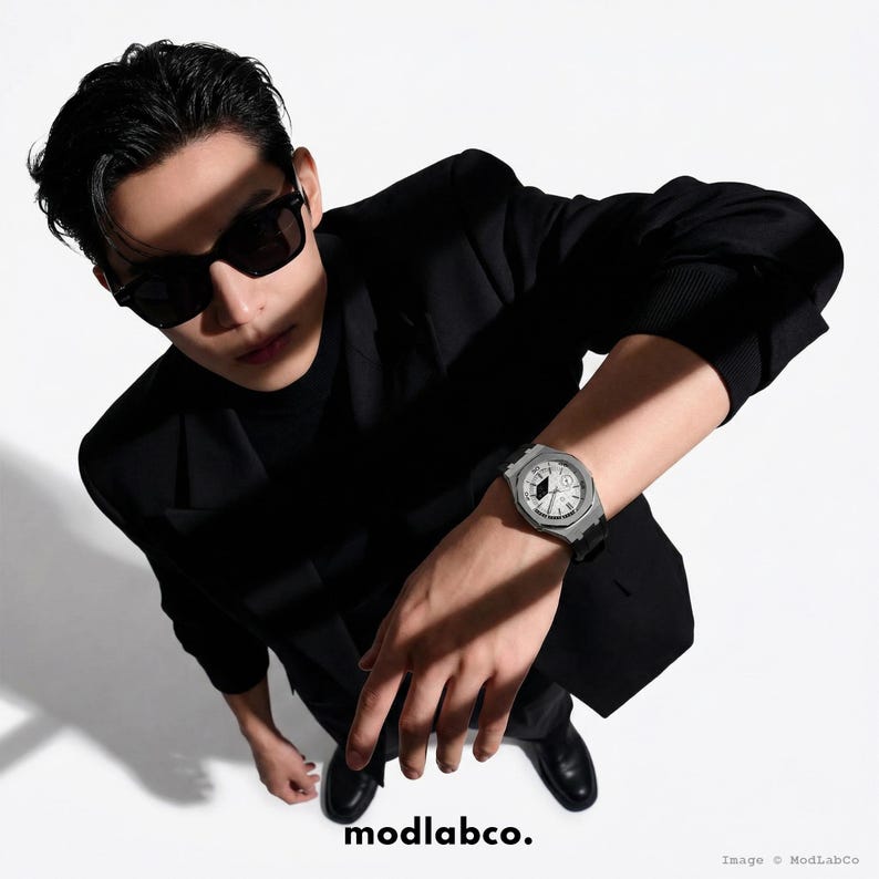May include: A silver wristwatch with a white face and black hands is displayed. The watch has a black band and is worn on a wrist. The image also shows a person wearing sunglasses and a black suit. The text "modlabco." is at the bottom.