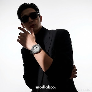 May include: A silver-toned wristwatch with a white face and black band is displayed. The watch is on a wrist, and the person is wearing a black blazer and sunglasses. The brand name "modlabco." is visible at the bottom of the image.
