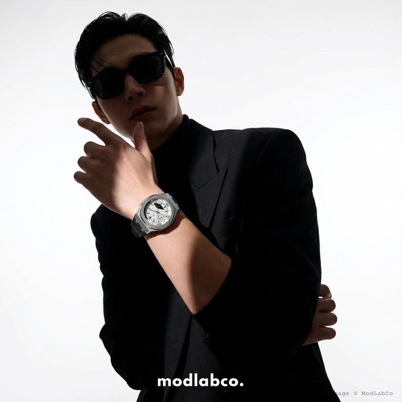 May include: A silver-toned wristwatch with a white face and black band is displayed. The watch is on a wrist, and the person is wearing a black blazer and sunglasses. The brand name "modlabco." is visible at the bottom of the image.