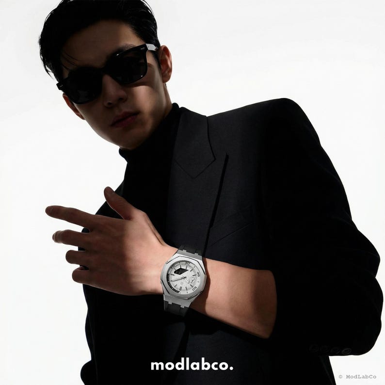 May include: A silver-toned wristwatch with a white face and black band is displayed. The watch is on a wrist, with a black suit and sunglasses in the background. The text "modlabco." is at the bottom.