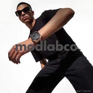 Op de afbeelding: Een zwart horloge met een roségouden kast en een zwarte wijzerplaat wordt om de pols van een persoon gedragen. De persoon draagt een zwart t-shirt en een zwarte jeans. De afbeelding bevat de tekst "modlabco."