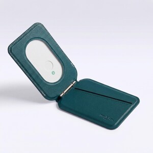 May include: Teal leather wallet, open to show a white card with a rounded rectangle cutout. The wallet has a hinged design with white stitching. The brand name is embossed on the bottom right.