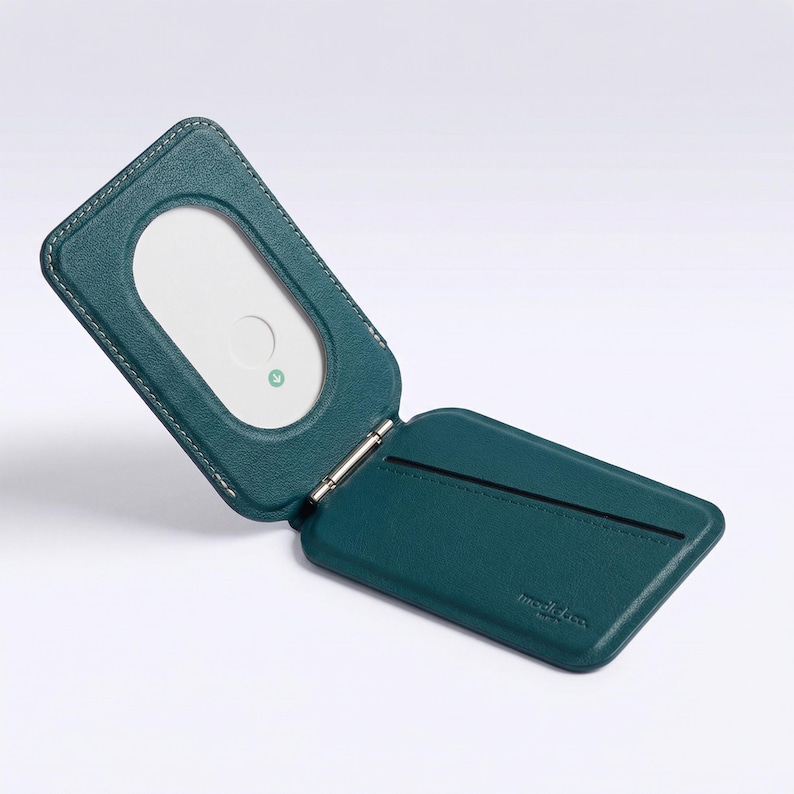 May include: Teal leather wallet, open to show a white card with a rounded rectangle cutout. The wallet has a hinged design with white stitching. The brand name is embossed on the bottom right.